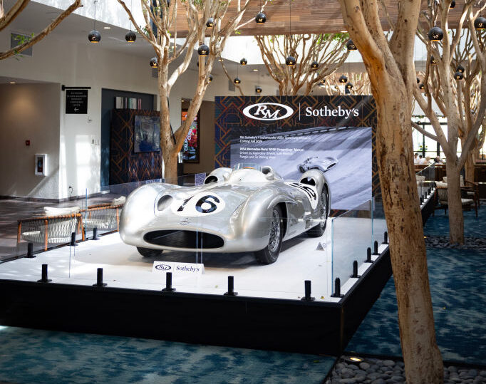 A vintage silver race car is displayed on a platform under a wooden pergola in a spacious gallery with tall trees and modern lighting. A Sotheby's banner is in the background.