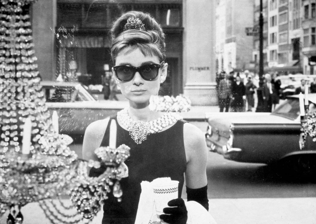 A stylish woman in a black dress, pearl necklace, and large sunglasses stands by a storefront window. She holds a paper bag and is surrounded by sparkling jewelry. A classic car and city street are visible in the background.