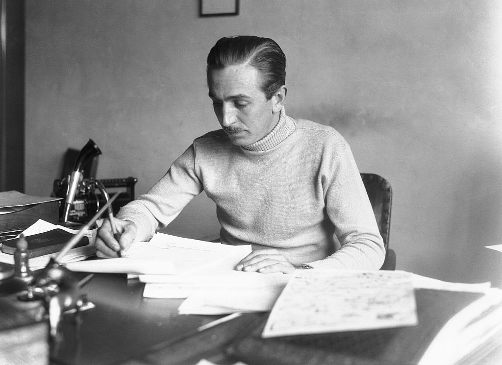 A man sitting at a desk, wearing a light-colored sweater, is focused on writing or drawing on papers spread out in front of him. The room appears to be an office setting with additional documents and a telephone visible on the desk.