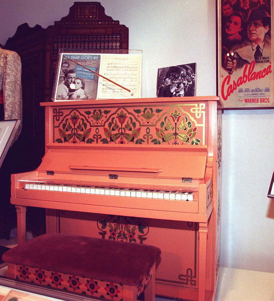 A vintage, ornately decorated upright piano with floral patterns. Sheet music and a framed photograph are displayed on top. A "Casablanca" movie poster is partially visible on the wall behind the piano.