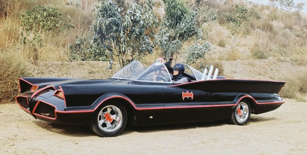 A black custom car with red accents, resembling a bat motif, drives on a dirt road surrounded by dry brush and hills. A person wearing a blue costume and mask is seated inside the vehicle.