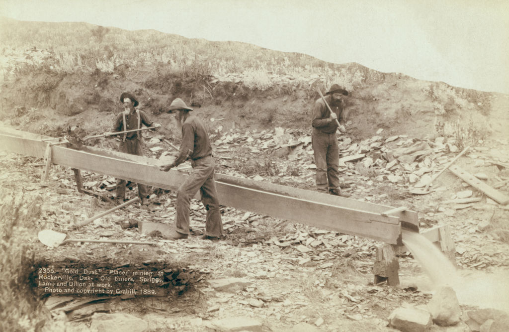 Three miners in 1889 work at a gold placer mine in Rockerville, Dakota. Two men use shovels to sift through dirt and rocks, while a third manages a sluice box, surrounded by a rugged, rocky landscape.