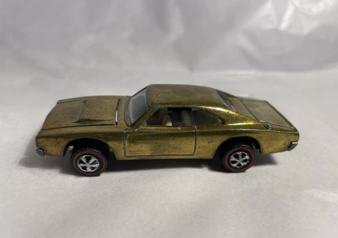 A vintage Hot Wheels toy car with a worn, metallic gold finish sits on a plain white surface. The model features a sporty design with detailed wheels and a sleek body, reflecting its classic style.