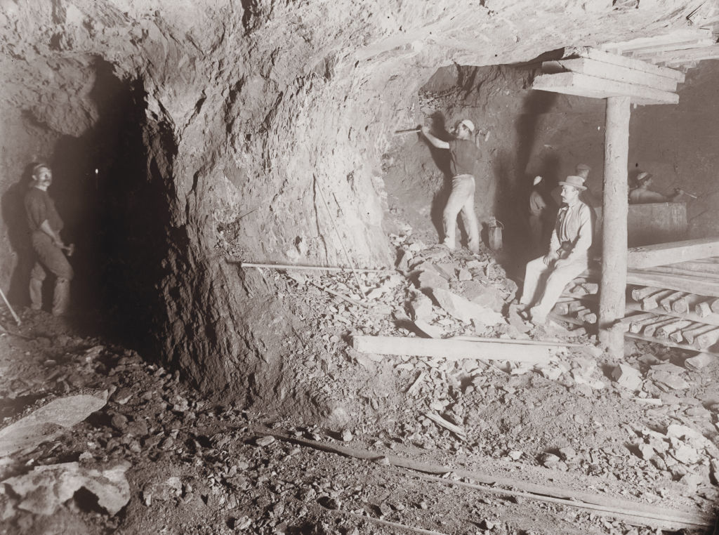 Miners working underground in a dimly lit tunnel, surrounded by rock walls. Three men are visible, two standing and one sitting on wooden supports. Tools and rubble are scattered on the ground. A rail track runs through the tunnel.