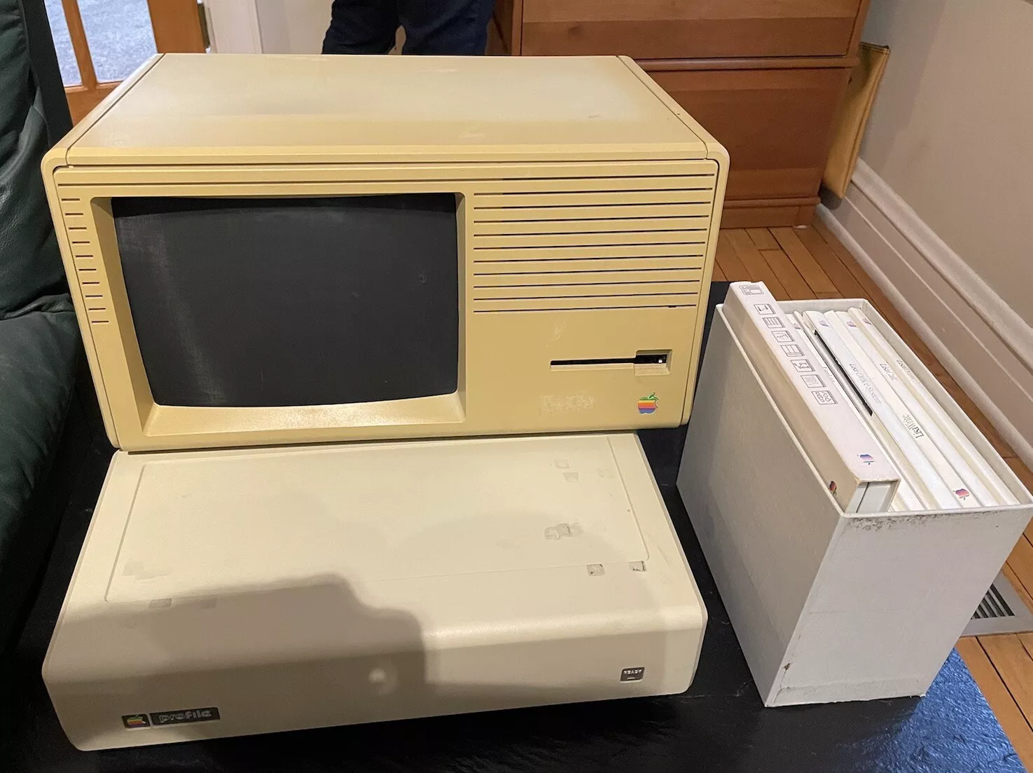 A vintage Apple Lisa computer with a monitor and a keyboard. To the right, there is a stack of white binders or manuals in a holder on a wooden floor. The equipment is placed on a black surface.