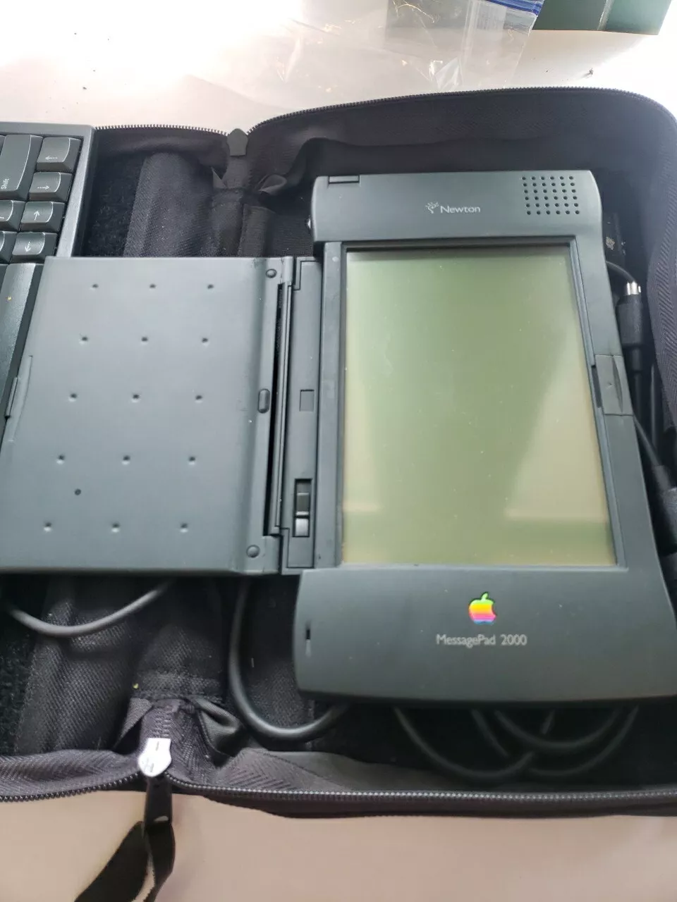 A Newton MessagePad 2000, an early PDA device with a stylus resting on its open screen, is placed next to a keyboard. The device is in a black protective case.