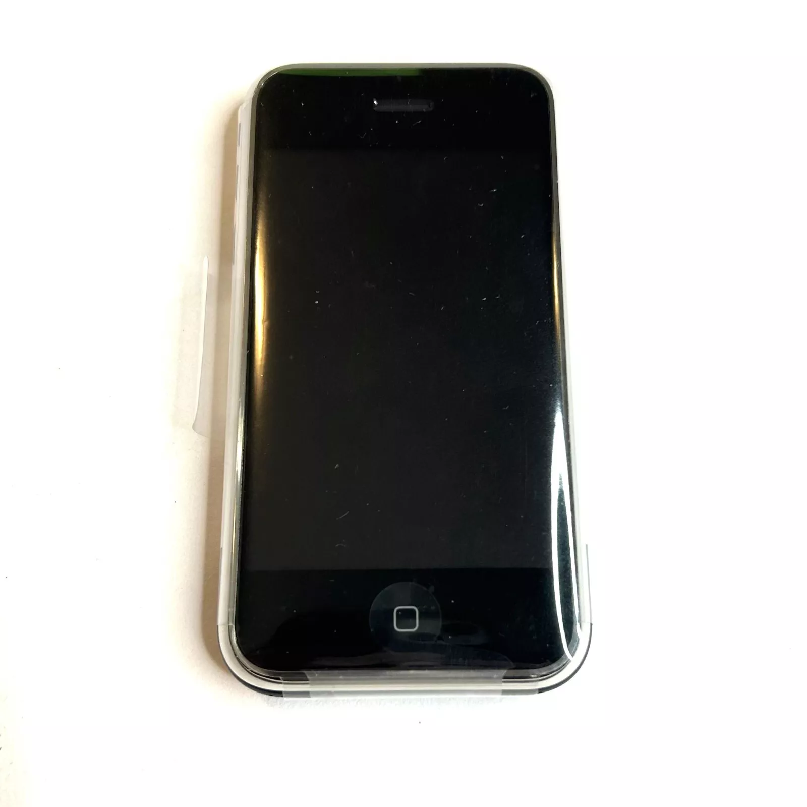 A black smartphone with a reflective screen and a home button at the bottom is resting on a white surface. It appears to be an older model and is encased in a clear protective cover.