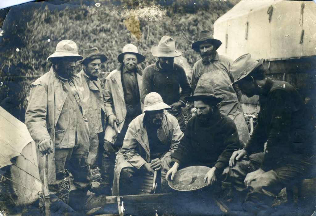 A vintage black and white photograph shows a group of nine men dressed in rugged outdoor clothing and hats. They appear to be gold prospectors gathered around a pan, engaged in discussion. A forested background and tents are visible.