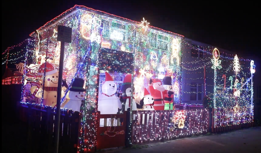 A house covered in colorful Christmas lights and decorations, featuring inflatable figures like snowmen, Santa Claus, and holiday-themed images. The lights illuminate the house and yard against the dark night sky.