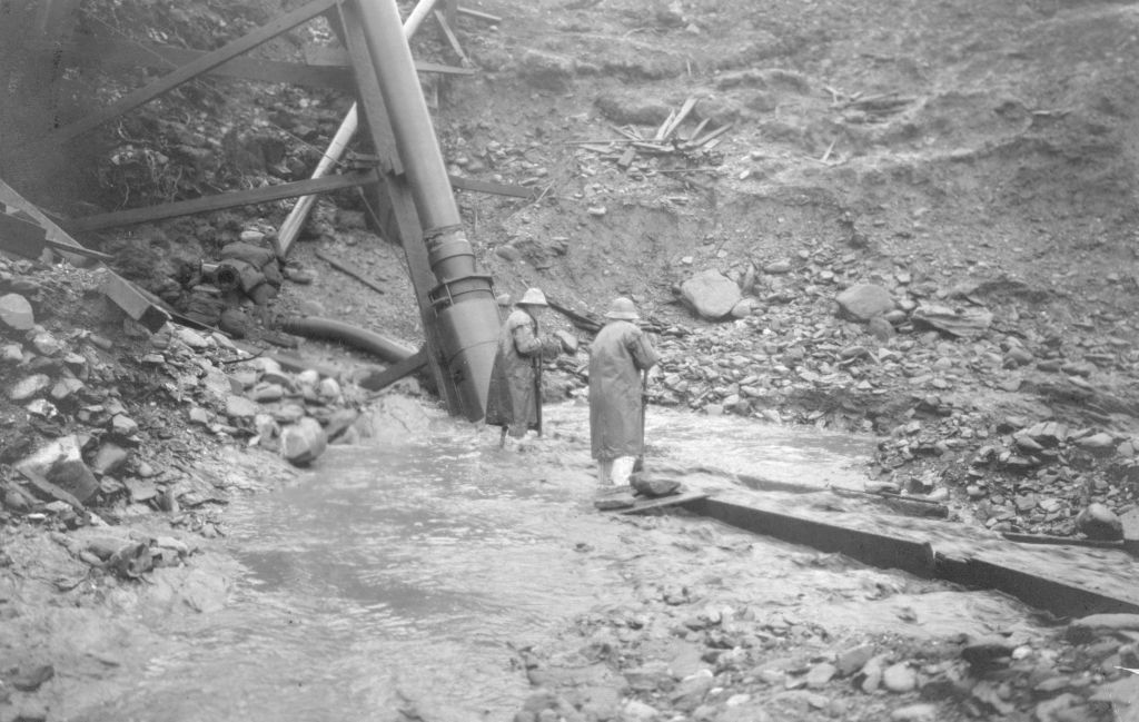 Black and white photo of two people in raincoats standing by a large pipe in a rocky, muddy area with flowing water. The scene suggests construction or mining activity.