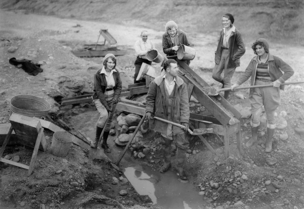 A group of people, including women and men, work together at a mining site. They are using shovels and a sluice to process materials. The scene appears to be from a past era, with everyone dressed in traditional mining attire.