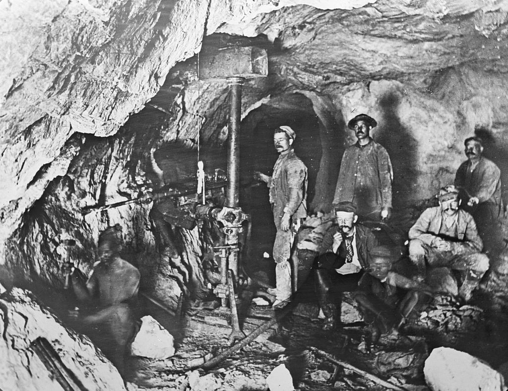 Black and white image of six miners working inside a cave. Some are standing, while others are sitting near mining equipment. The surrounding rock walls are rough, indicating an active mining operation. The scene is dimly lit, revealing labor-intensive conditions.