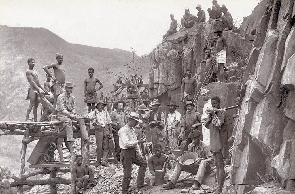 A black-and-white historical photograph shows a group of men working on a mountainside, likely involved in construction or mining. Some are using tools, while others stand or sit on the rocky terrain. The area appears rugged and mountainous.