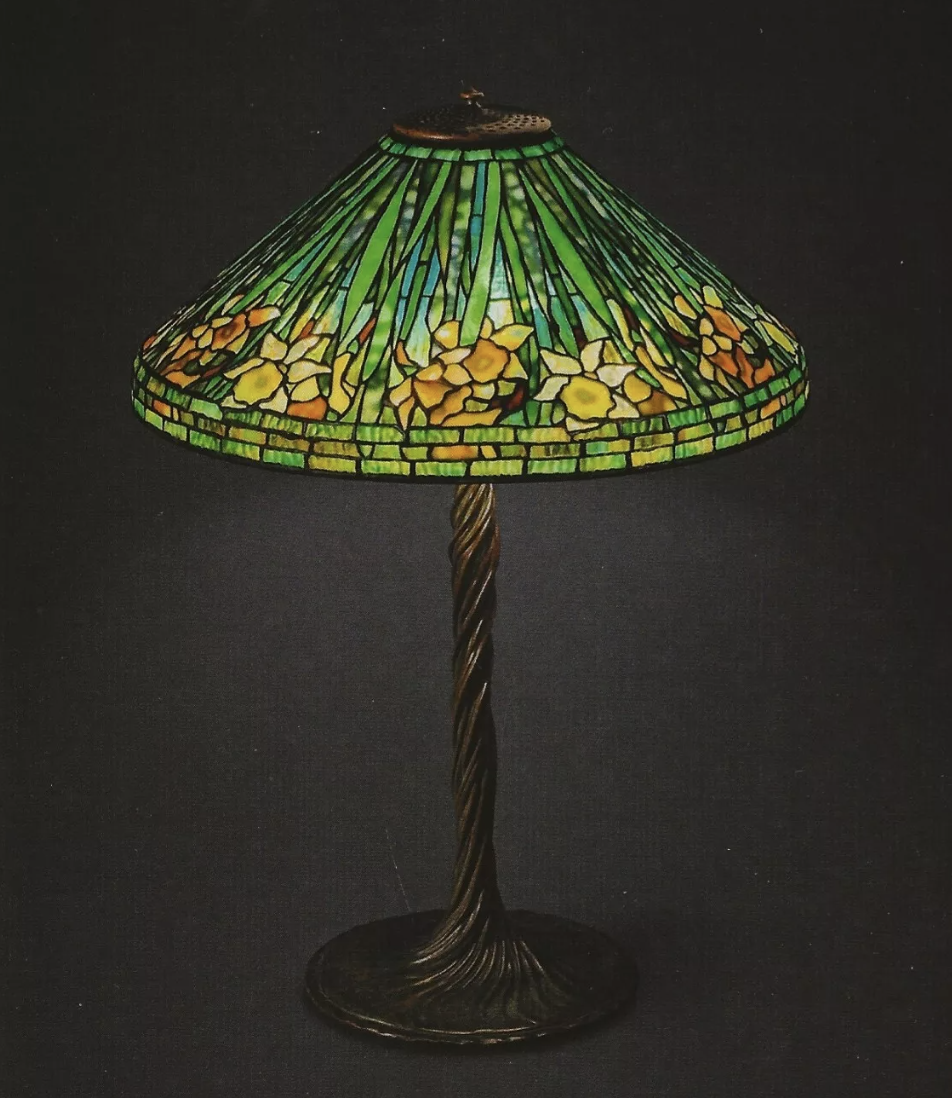 A stained glass table lamp with a floral design, this expensive Tiffany lamp features a predominantly green lampshade adorned with yellow and orange flowers. The base is intricately twisted, creating a striking contrast against the dark background.