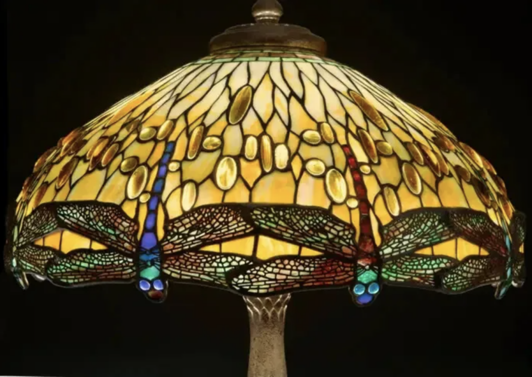 A stained glass lamp with a dome-shaped shade, boasting intricate dragonfly designs in vibrant colors and patterns. Reminiscent of expensive Tiffany lamps, the glass includes shades of yellow, green, and hints of blue and red, complemented by a bronze base.