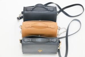 Three handbags, reminiscent of vintage Coach bags, are arranged vertically on a white surface. In dark gray, light brown, and black with long straps and a minimalist design, the black one stands out with its chain strap and small clasp closure.