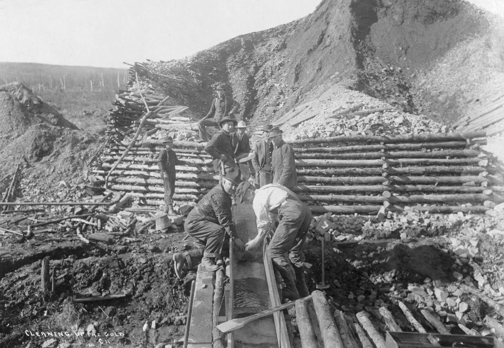 Workers in early 20th-century attire clean gold at a mining site. They stand and kneel on wooden structures amidst piles of rocks and earth. The background shows a large, earthy mound under a clear sky.