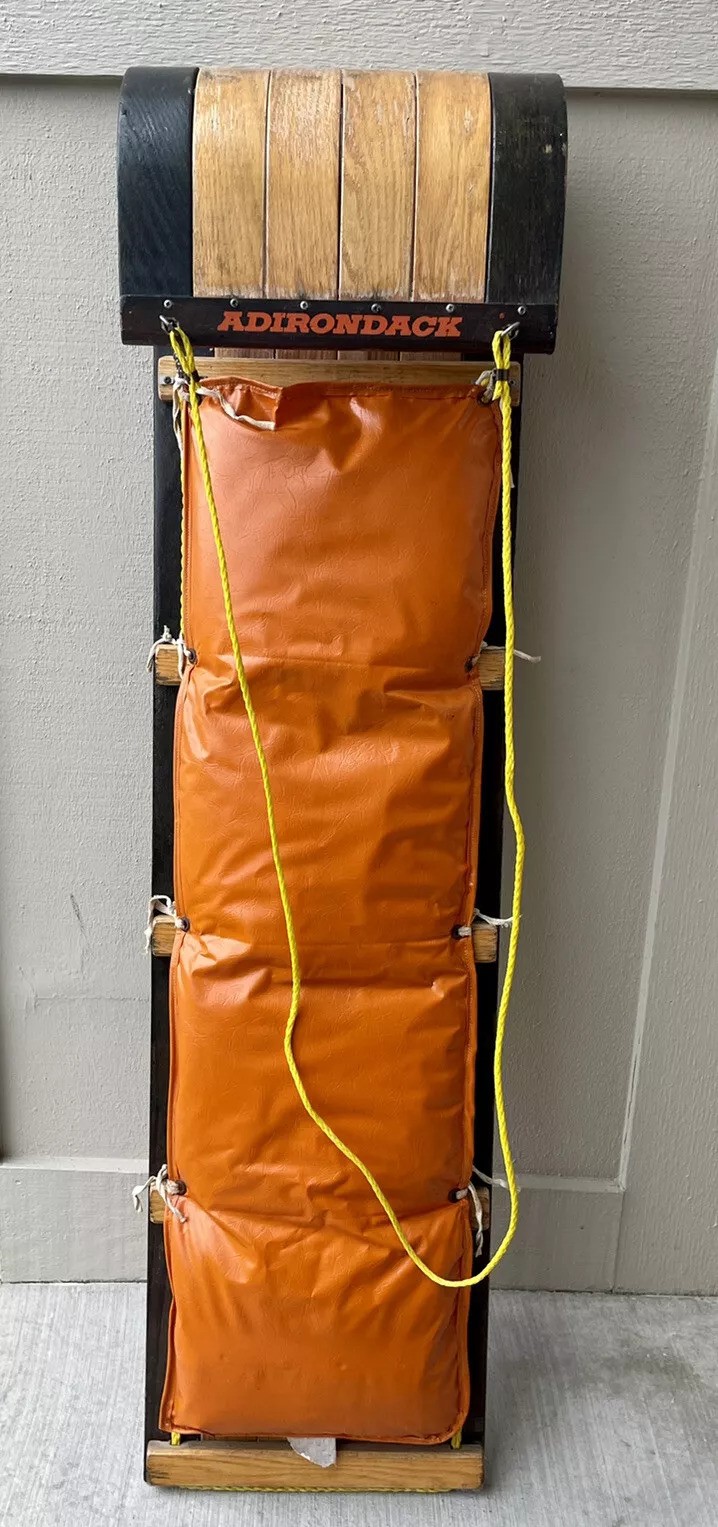 A vintage Adirondack toboggan with a wooden curved top, four orange padded sections, and a yellow rope handle hanging on a wall.