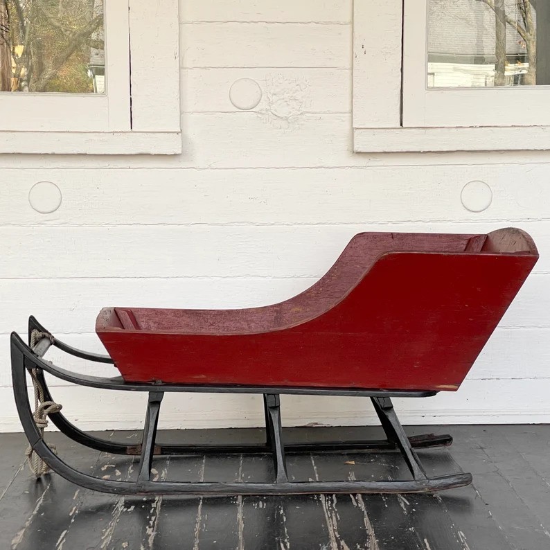 A vintage red wooden sleigh with black metal runners sits on a dark wooden floor. The sleigh is positioned in front of a white, textured wall featuring two circular decorations and a window with visible reflections.