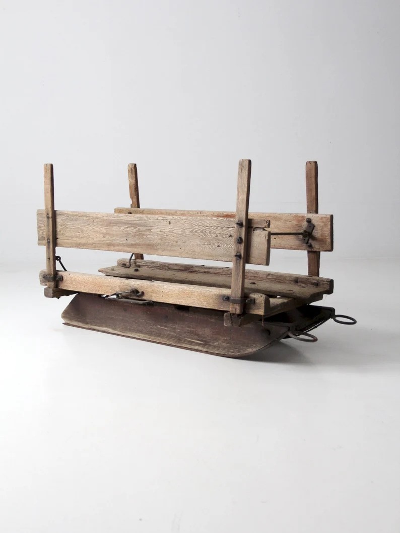 Wooden sled with a simple, rustic design. It features vertical posts and horizontal slats on a flat wooden base, with metal runners underneath. The sled is set against a plain, light-colored background.