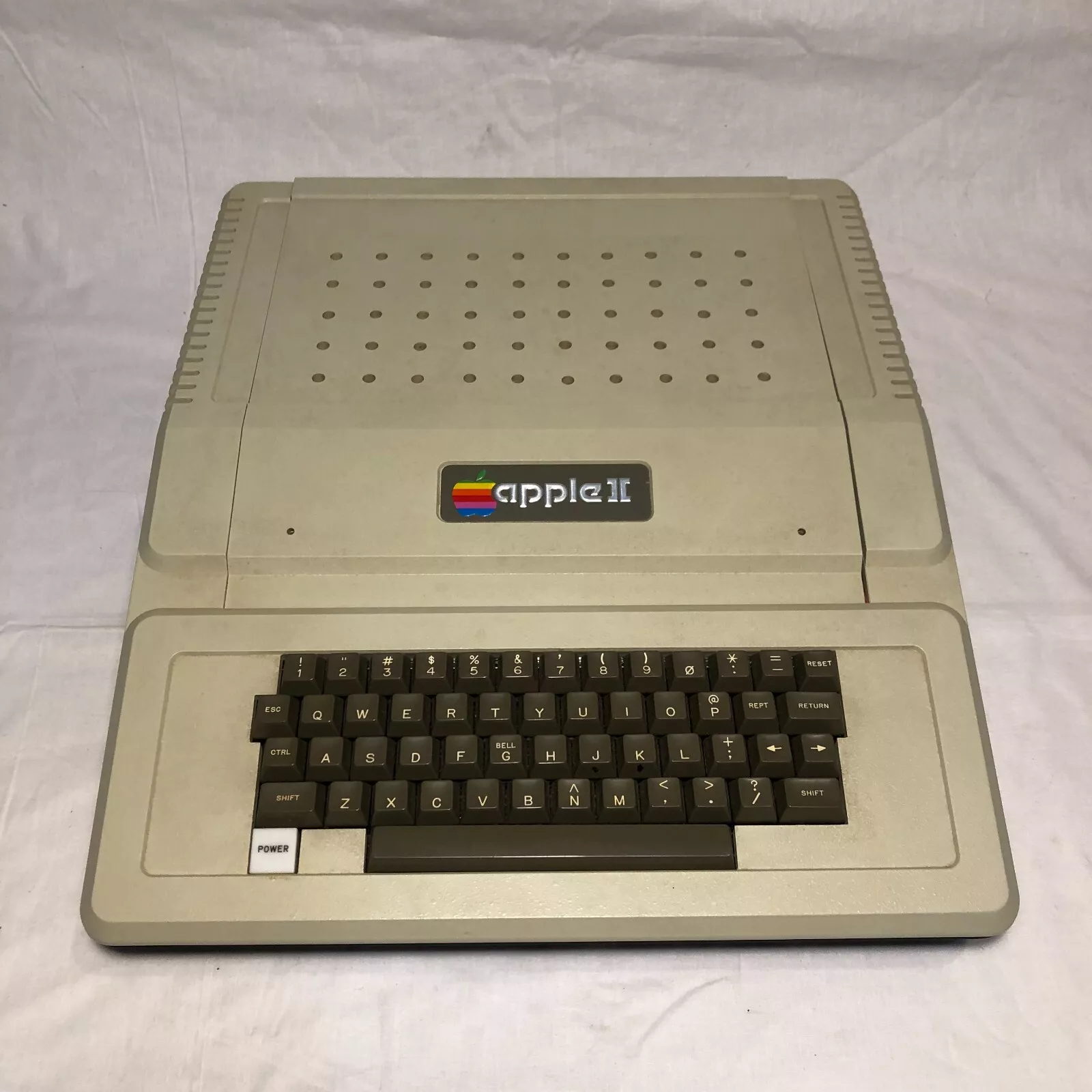 A vintage Apple II computer with a beige casing, featuring a built-in keyboard. The Apple II logo with a rainbow design is visible above the keyboard. The computer is placed on a plain white surface.