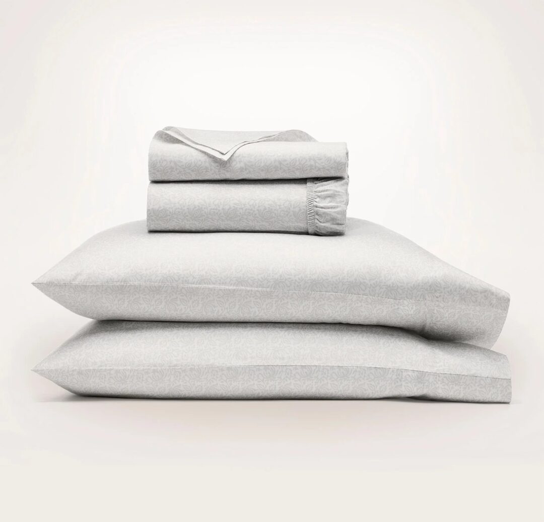 A neatly stacked set of light gray bed linens. Two pillows rest at the bottom, topped with a folded fitted sheet, a flat sheet, and a pillowcase. The fabric has a subtle, textured pattern. The background is plain white.