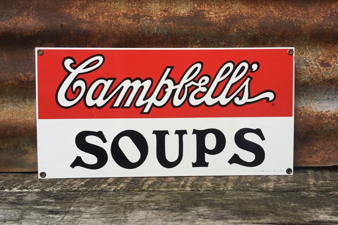 A vintage Campbell's Soups sign with a red and white background is displayed against a rusty metal surface. The word "Campbell's" is written in a cursive style, while "Soups" is in bold block letters. The sign has a weathered appearance.