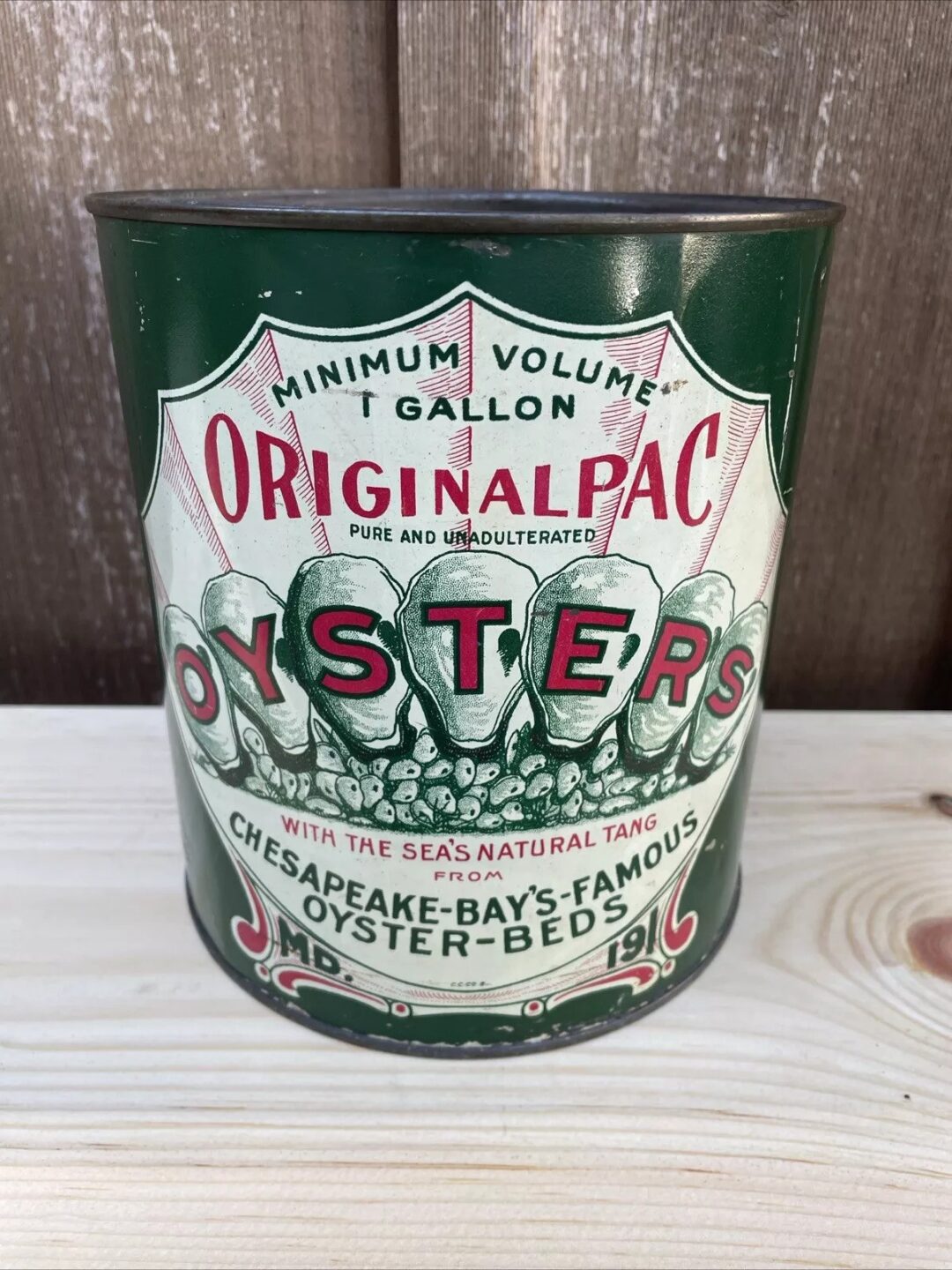 A vintage green and white can labeled "Original Pac Oysters," with red text and images of oysters. The label mentions "Minimum Volume 1 Gallon" and "Chesapeake Bay's Famous Oyster Beds." The can is placed on a wooden surface.