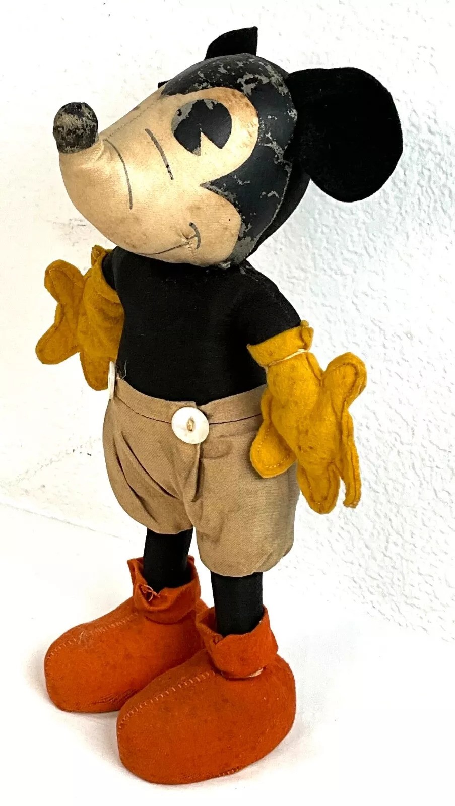 A vintage cartoon character toy stands with large round ears, black body, cream shorts, yellow gloves, and red shoes, showing wear and age. It's positioned against a white background.