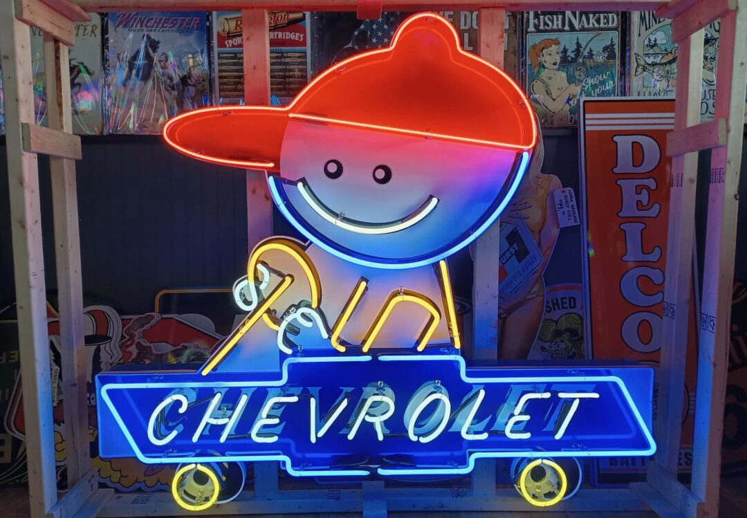 A vibrant neon sign featuring a cheerful figure in a red cap riding a blue and yellow skateboard, with the word "Chevrolet" displayed below. The background includes vintage posters and signs in a wooden frame.