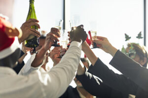 A lively party unfolds as a group of people clink champagne glasses in a celebratory toast. Some don festive hats, while bright, natural light fills the space, highlighting the joyful atmosphere.