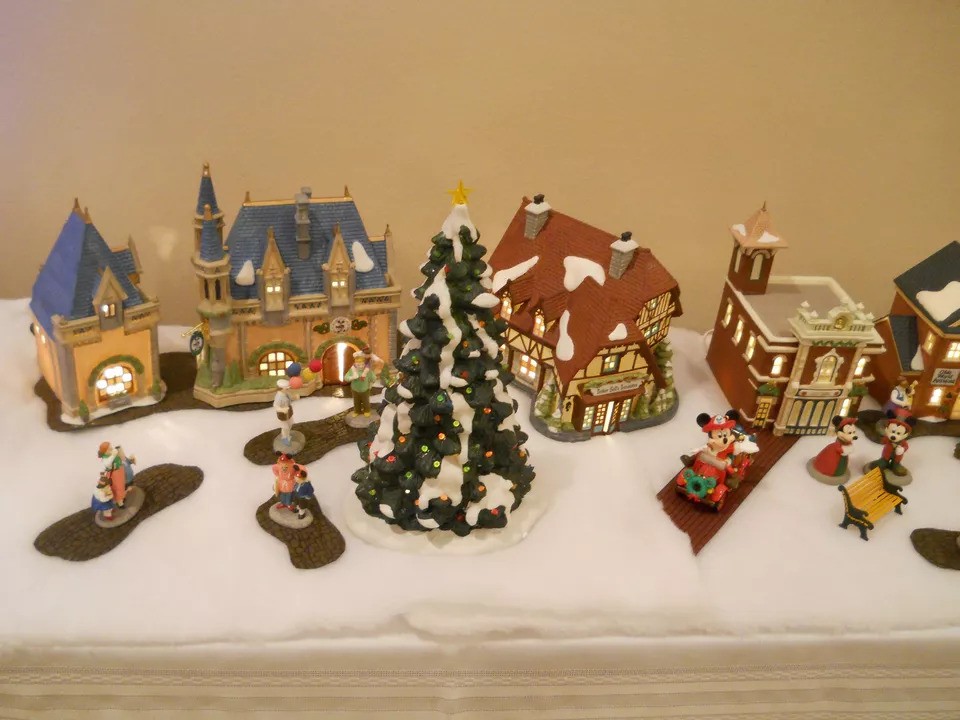 A charming miniature Christmas village display with a snow-covered base. It features a central Christmas tree, various colorful buildings, and festive figurines, including a couple in a sleigh.