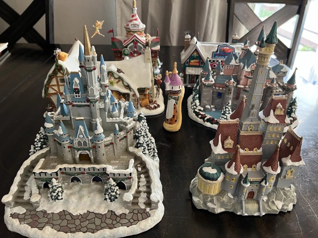 A collection of detailed miniature castles and holiday-themed village buildings are displayed on a dark table. The castles are adorned with snow, and one features a small golden figure flying above it.