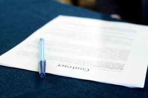 A pen rests on a partially readable contract nestled on a blue fabric surface. The document, slightly angled, prominently displays the word "contracts" at the bottom.