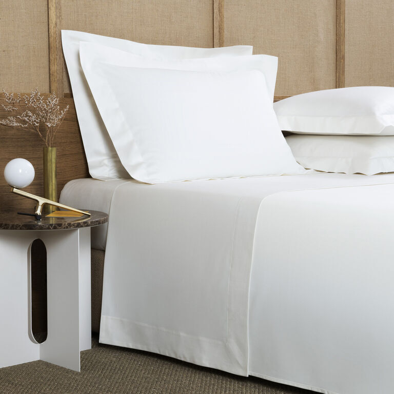 A neatly made bed with white sheets and pillowcases, set against a wooden headboard. A white side table on the left holds a small lamp and dried flowers. The floor is carpeted in a neutral tone.