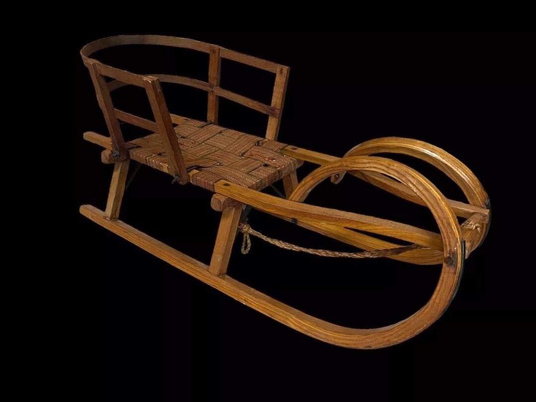 A vintage wooden sled with a curved design and a woven seat, featuring slatted sides and a rope attached to the front for pulling, set against a black background.