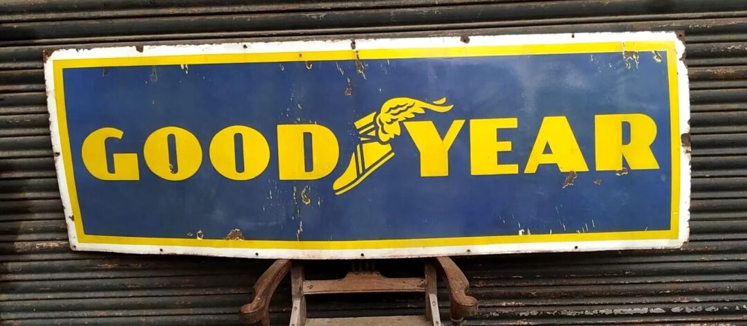 An old, weathered Goodyear sign with a blue background and yellow lettering is propped against a corrugated metal shutter. The sign features the iconic winged foot logo.