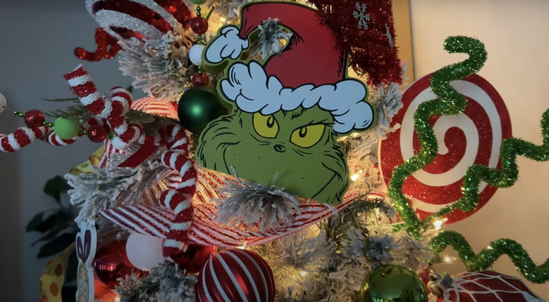 A festive Christmas tree adorned with a large Grinch face decoration wearing a Santa hat. The tree is decorated with red and white striped ribbons, sparkling green and red ornaments, and twinkling lights, creating a whimsical holiday display.