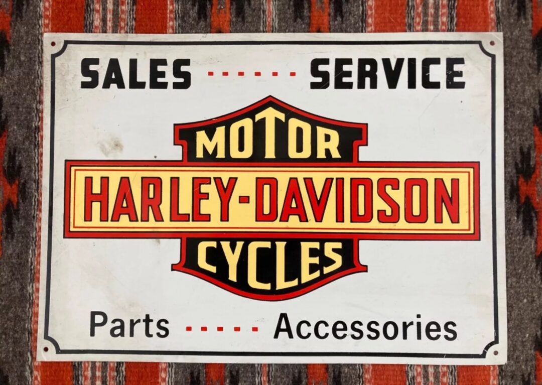 Vintage Harley-Davidson sign with red, yellow, and black lettering on a patterned background. Text reads "Sales Service Motor Harley-Davidson Cycles Parts Accessories.