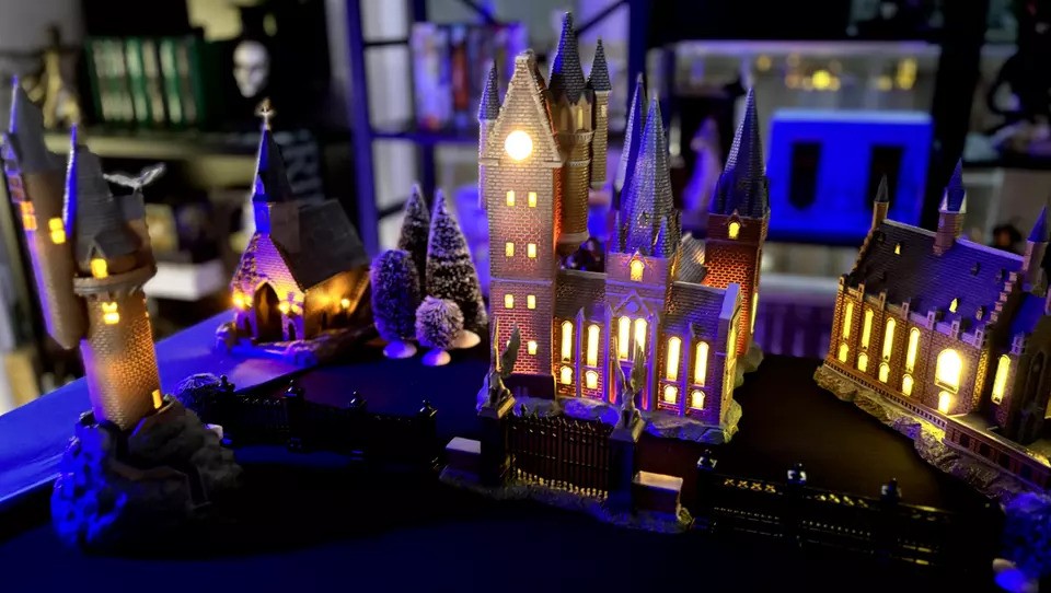 A detailed, illuminated model of a magical castle at night, featuring tall spires and glowing windows. Surrounded by trees, with a dark background highlighting the intricate architecture.