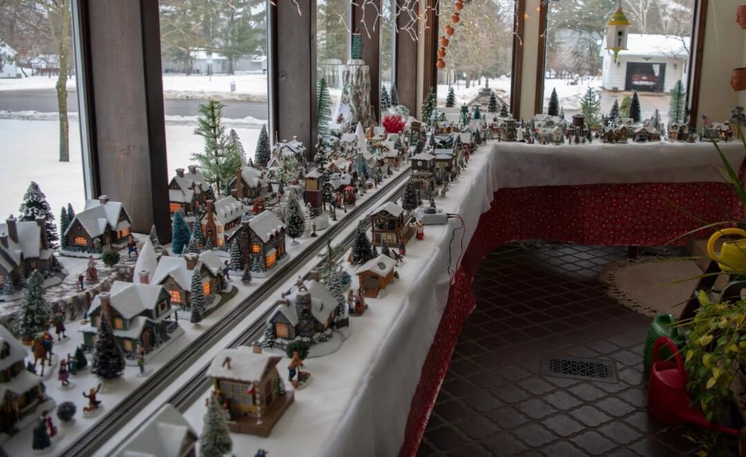 A festive indoor display of a miniature winter village, featuring snow-covered houses, tiny evergreen trees, and twinkling lights. A model train is set on tracks winding through the village. Snowy landscape is visible through the window.