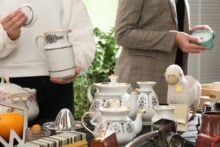 Two people browsing items at a flea market. One holds a white ceramic pitcher and lid, while the other examines a blue alarm clock. The table displays various vintage teapots, a plush sheep, and other miscellaneous items.