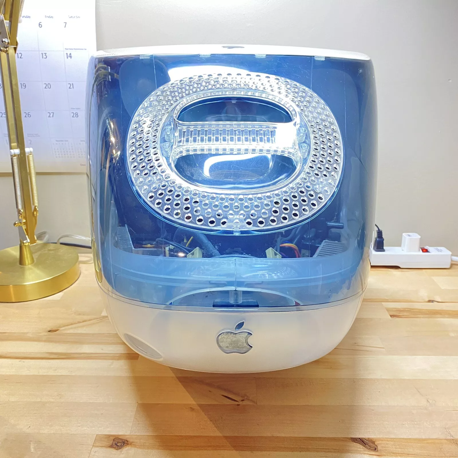 A vintage Apple iMac G3 computer with a translucent blue casing is on a wooden desk. A calendar and a gold desk lamp are visible in the background.