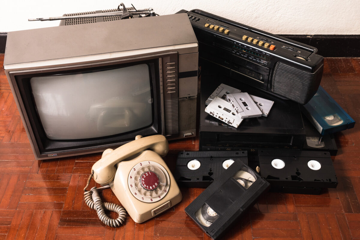 A vintage collection featuring a CRT television, rotary dial telephone, cassette tapes, VHS tapes, and a stereo system with a cassette player, all arranged on a wooden floor.