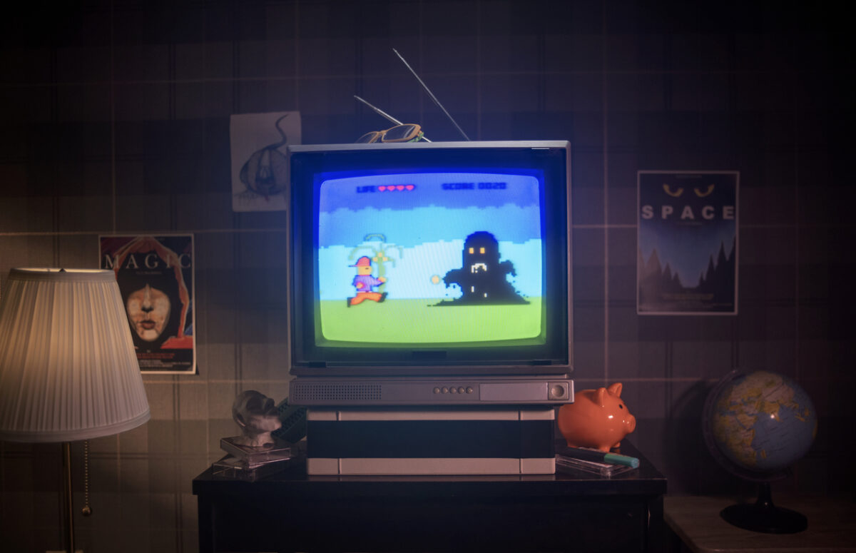 A vintage CRT television displays a retro video game with a character facing a dark figure. The room has a warm lamp, posters on the walls, a globe, and a piggy bank on a shelf, creating a nostalgic ambiance.