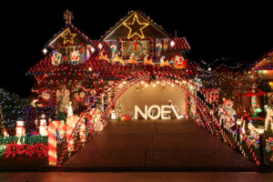 A house decorated with bright, colorful Christmas lights at night. The display features a large star on the rooftop, multiple festive figures, and the word "NOEL" in illuminated letters on the driveway. A bridge of lights arches over the entrance.