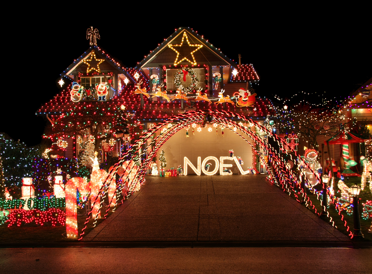 A house decorated with bright, colorful Christmas lights at night. The display features a large star on the rooftop, multiple festive figures, and the word "NOEL" in illuminated letters on the driveway. A bridge of lights arches over the entrance.