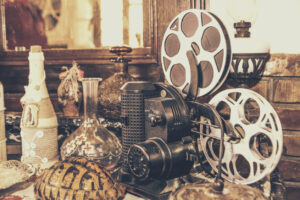 A vintage film projector surrounded by various decorative objects, including a bottle wrapped in rope, glass containers, and a patterned wooden item. The setting appears to be an antique or retro-themed room.