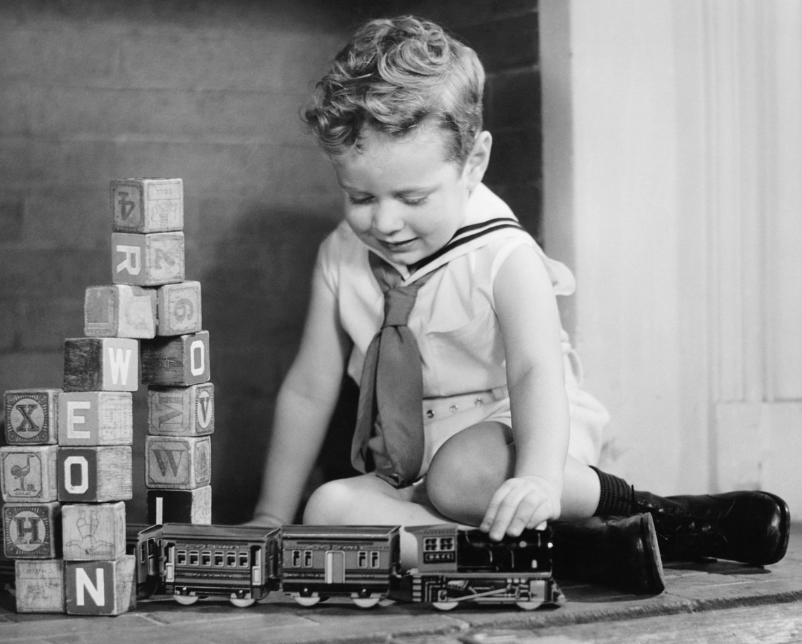 A young child wearing a sailor outfit plays with a toy train on the floor. Wooden blocks are stacked nearby. The child appears focused and is in a room with a brick wall background.