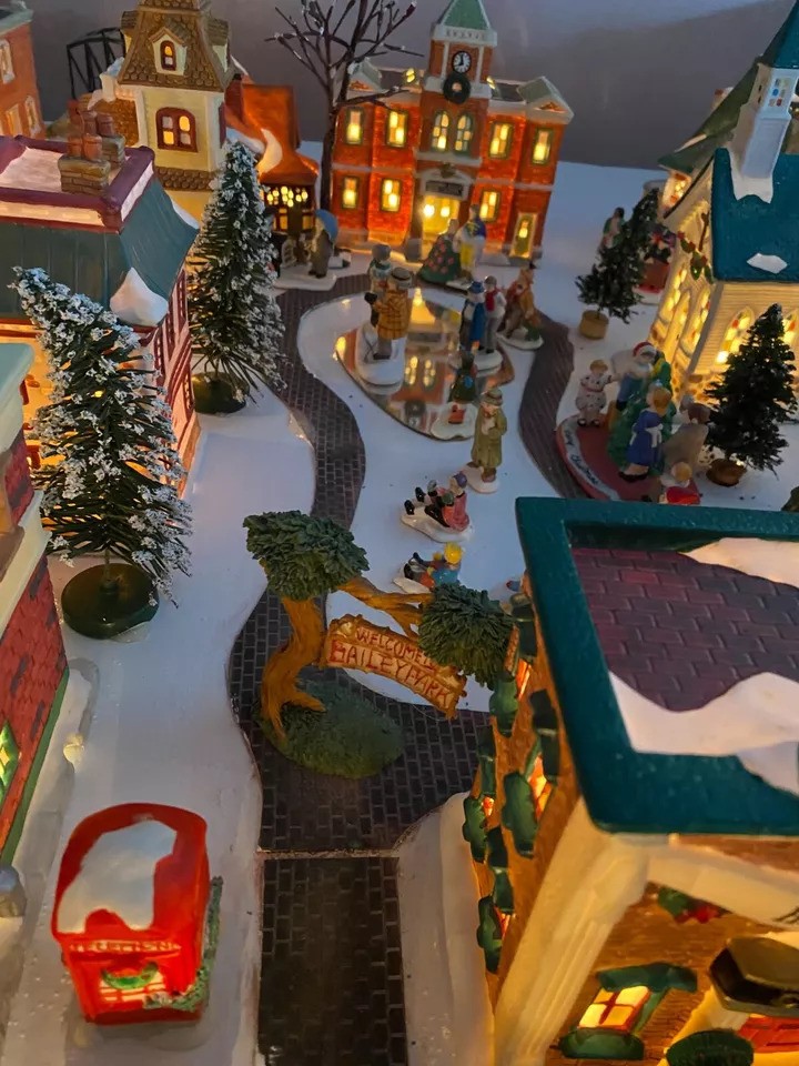 A festive miniature village scene with snow-covered buildings and trees, lit by warm lights. Tiny figures gather around a central square. A small red phone booth and a sign reading "Welcome to Mistletoe Valley" are visible in the foreground.